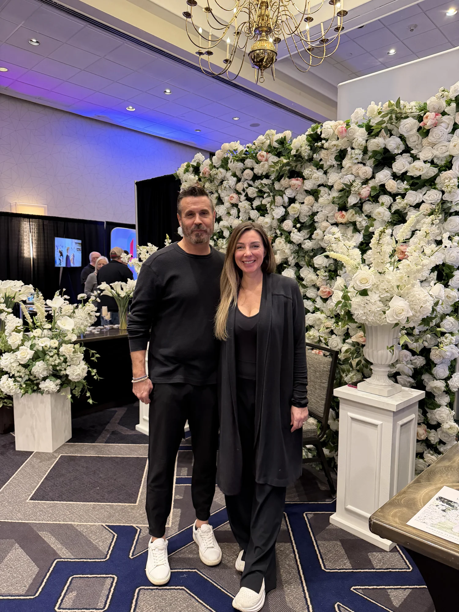 A couple posing in front of a lush white and blush floral wall at an Indy Engaged showcase