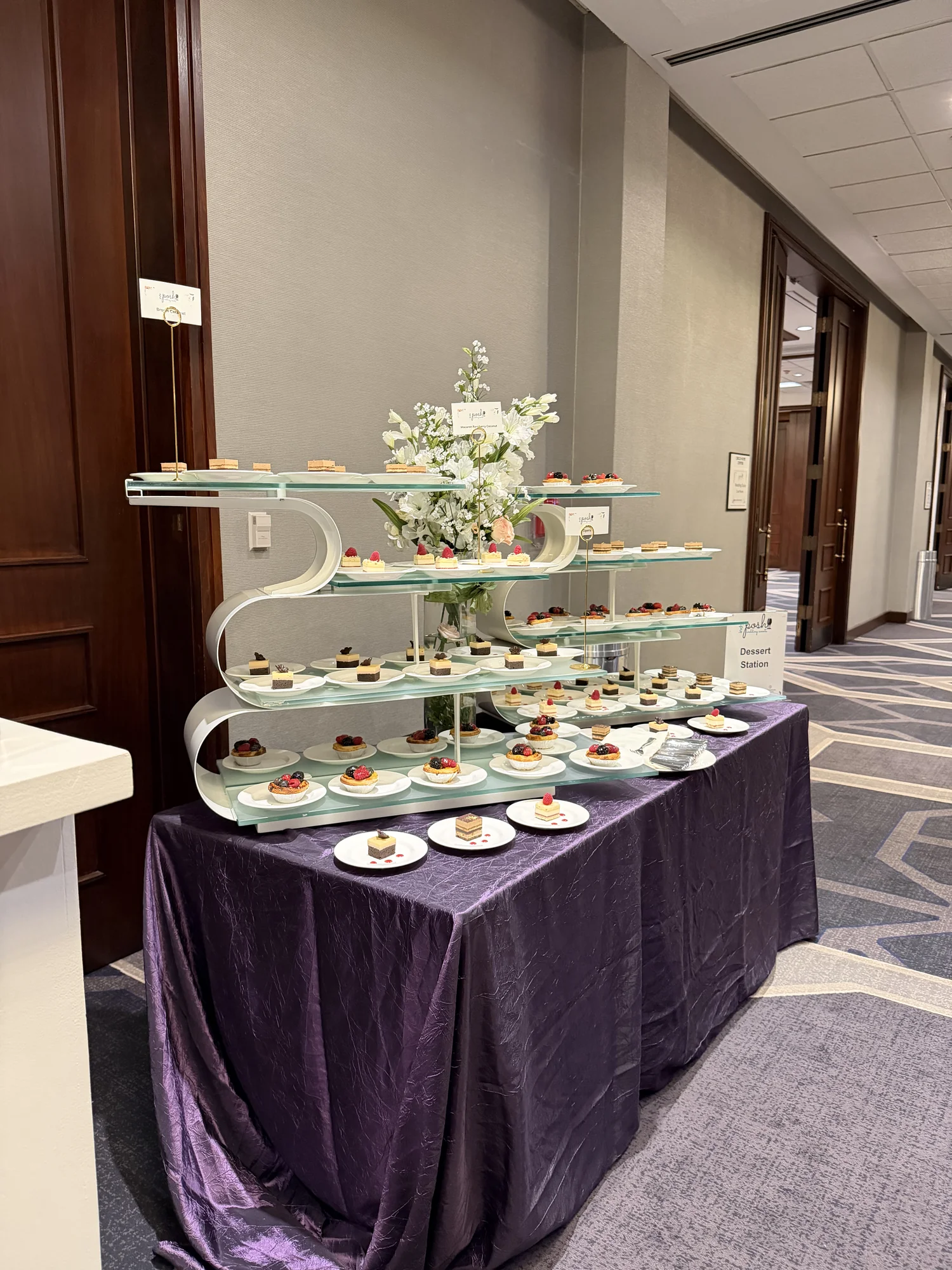 Tiered glass dessert display with miniature cakes and white florals on a purple linen