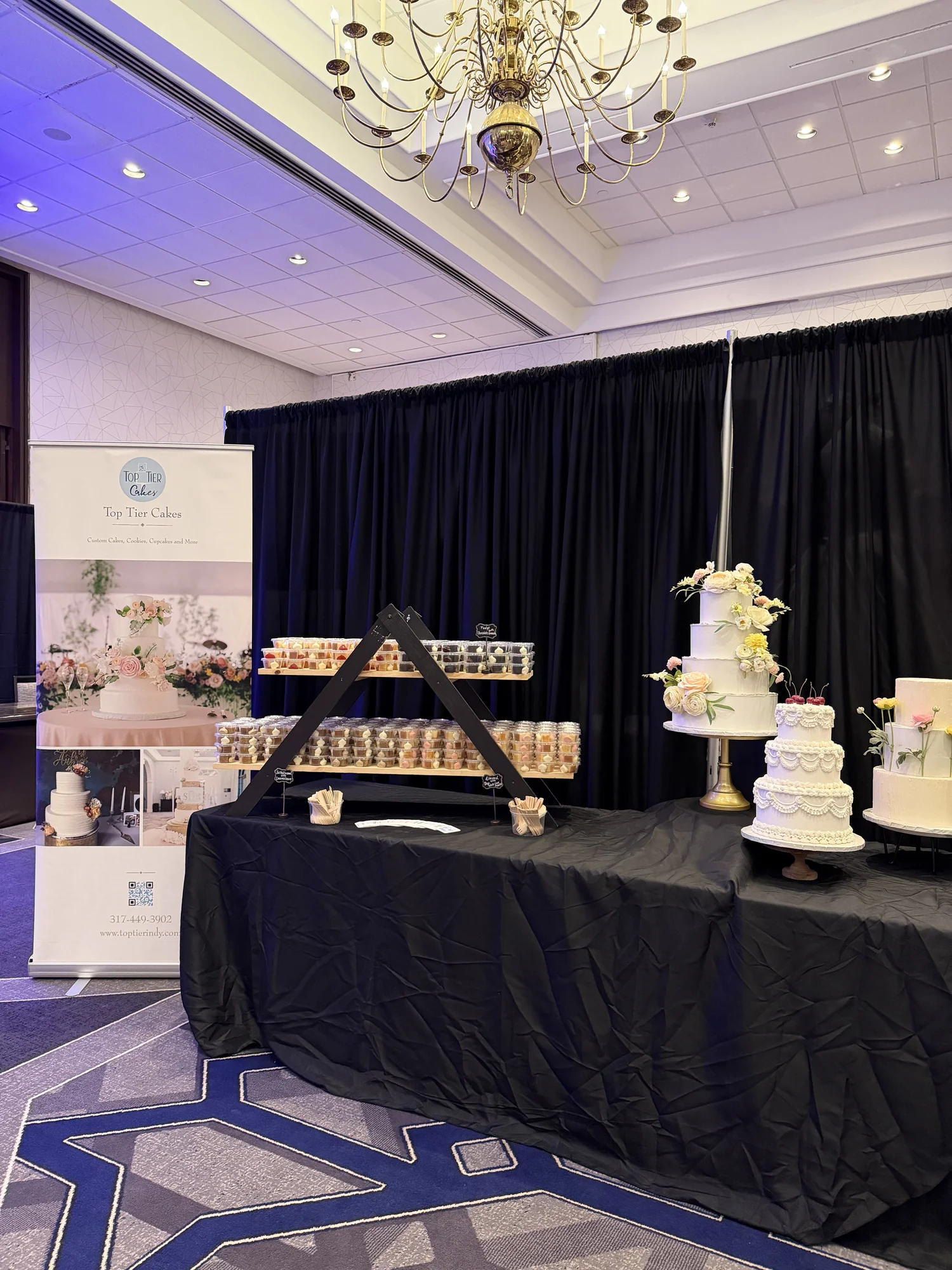 Top Tier Cakes booth with a tiered white wedding cake and cupcake display